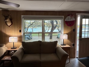 A cozy room with a wooden wall, a large window with a view of trees and a dock, a sofa, two lamps, and a door with a sign. - Lake Travis waterfront escape that sleeps 16 (Spicewood)