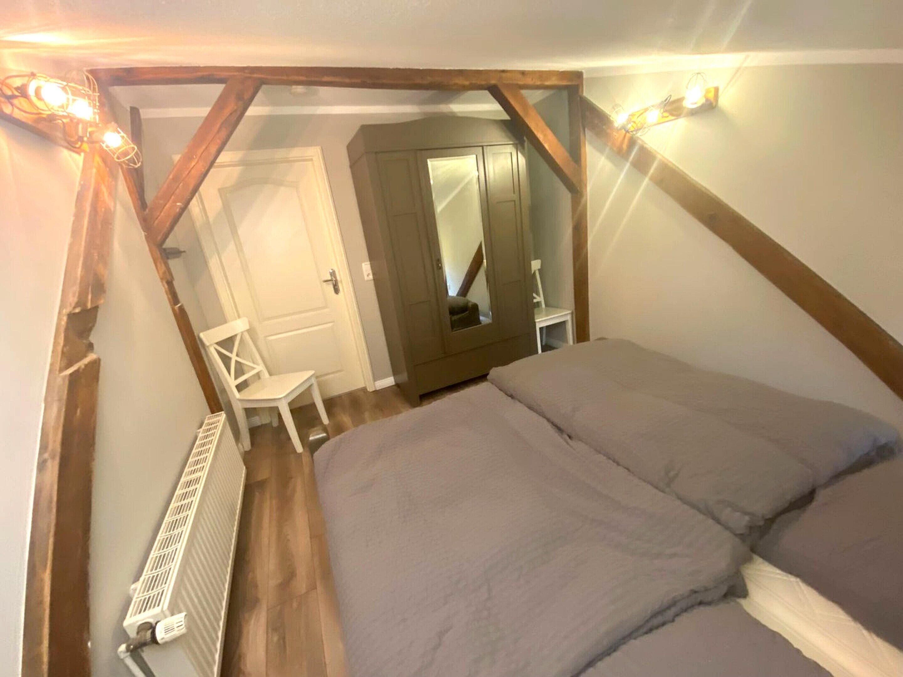 Ein Schlafzimmer mit schrägen Wänden, Holzbalken, einem Bett, einem Sessel, einem Kleiderschrank und einer Heizung.
