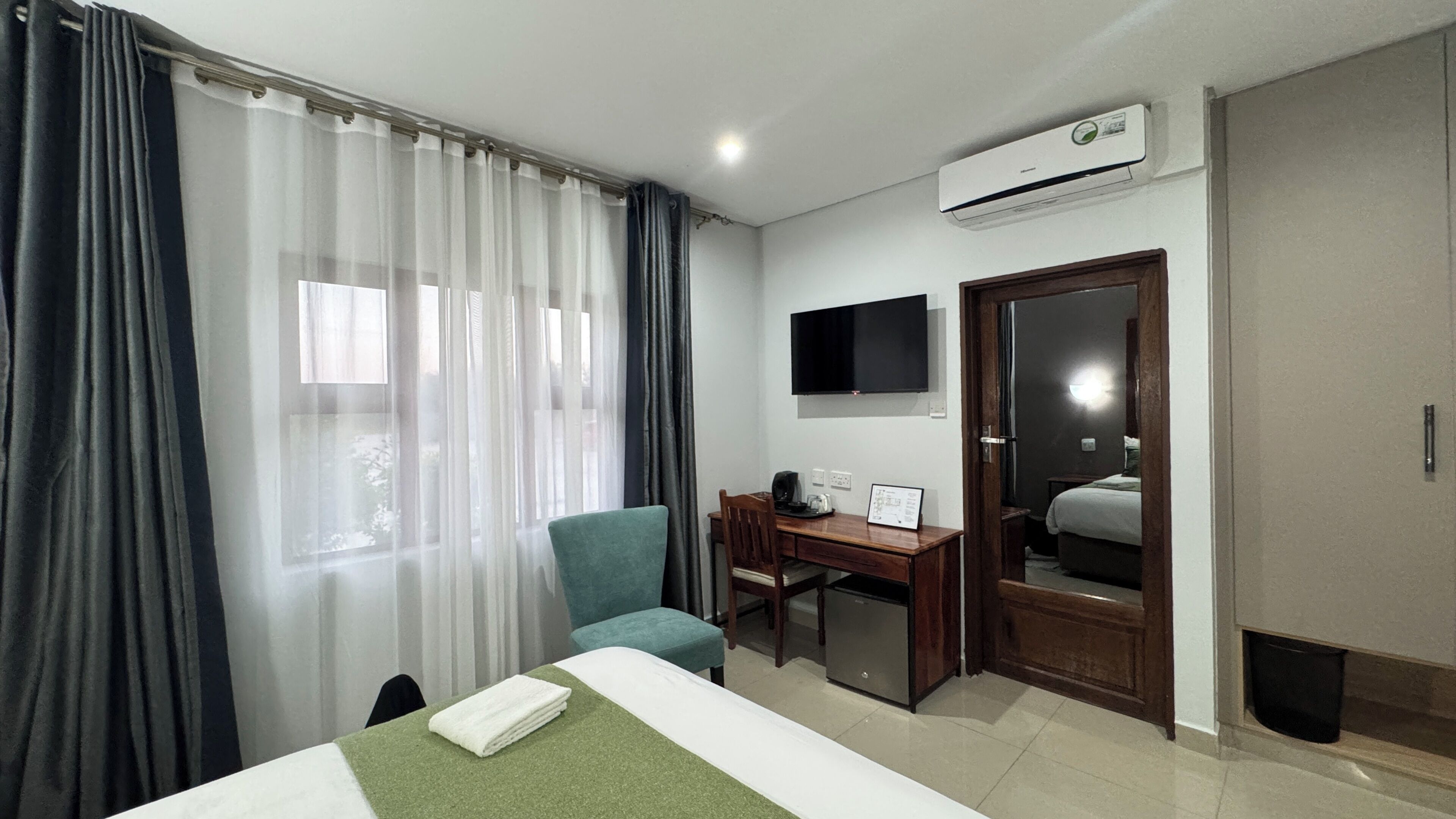 A hotel room with a bed, a desk with a chair, a flat-screen TV, and an air conditioning unit.