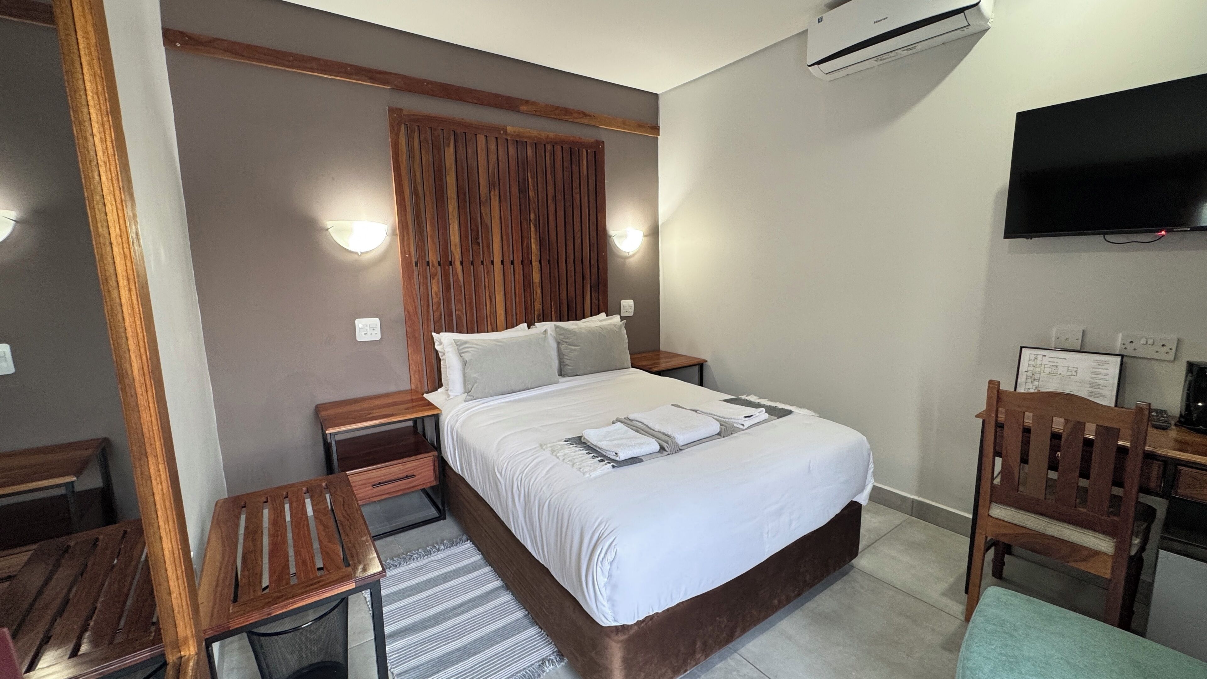 A bedroom with a bed, bedside tables, a desk, a chair, an air conditioning unit, and a television.