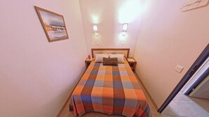 A small, neatly arranged bedroom with a checkered bedspread, a nightstand, a bedside table, and a wall-mounted light fixture. - Oceanfront Escape for 7 in Audierne (Audierne)