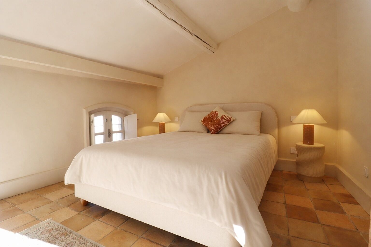 Um quarto com uma cama grande, duas luminárias de cabeceira, uma janela pequena e uma porta.