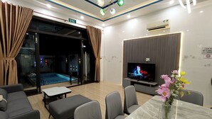 A modern living room with a TV, seating, a dining table, and a view of a pool. - Aroma-Pool-Breakfast-Man Thai beach (Da Nang)