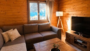 Ein gemütliches Blockhaus-Interieur mit Holzbalkendecke, einer Couch, einem Couchtisch mit Pflanze, einem Fernseher und einem Fenster mit Vorhängen.