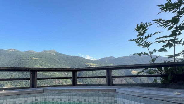 Ein klares Schwimmbecken mit Fliesenrand, umgeben von einer Holzdielenveranda und einem Metallgeländer, mit Blick auf eine Berglandschaft.
