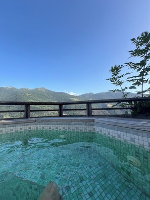 Une piscine aux eaux limpides, bordée de carreaux, entourée d’une terrasse en bois et d’une rambarde en métal, d’où l’on peut admirer un paysage montagneux.