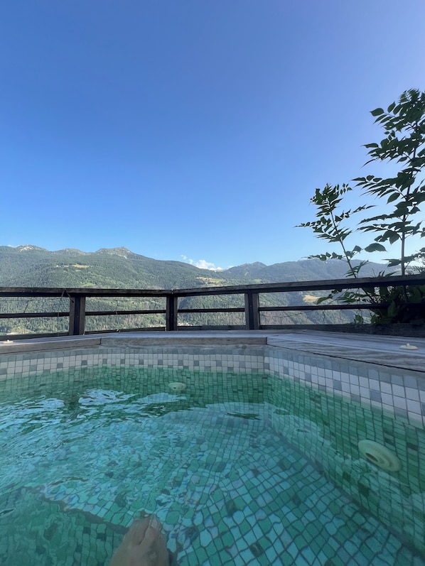A clear pool with a tiled edge, surrounded by a wooden deck and a metal railing, overlooking a mountainous landscape.