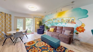 Una sala de estar moderna con un sofá, un sillón y un reposapiés, una mesa de comedor con sillas, y un mural grande en la pared.