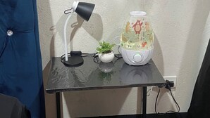 Una habitación pequeña con un escritorio, una lámpara, una planta, una fuente de agua y una silla.