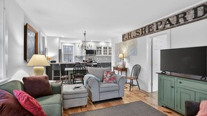 A living room with a sofa, ottoman, and a television on a cabinet. There is a kitchen in the background with white cabinets and a chandelier. - Forest Backdrop Midcoast Maine Quiet Escape 3 Bedroom Home (Dresden)