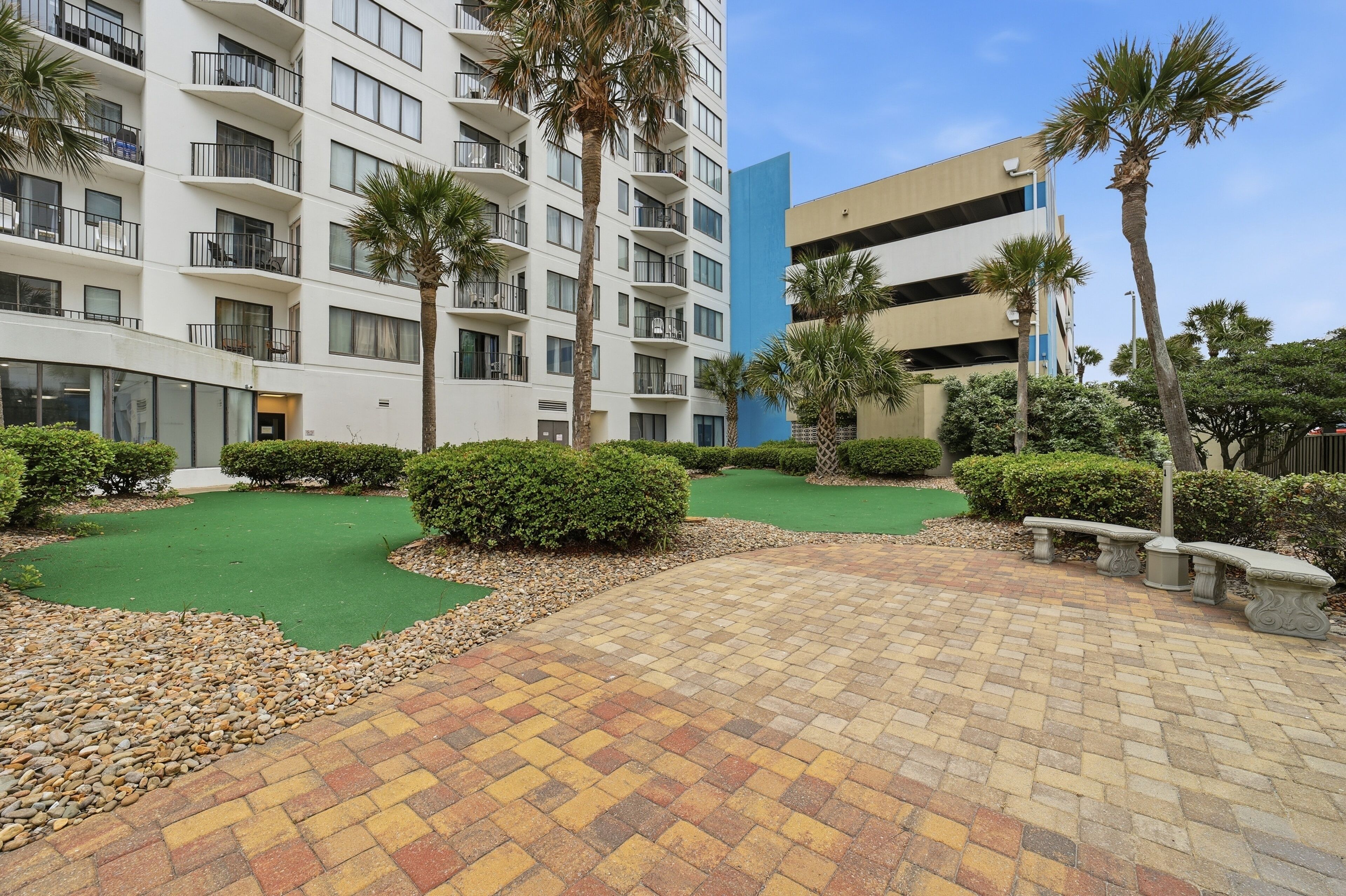 A courtyard with a green artificial turf, paved walkway, palm trees, and a multi-story building with balconies.