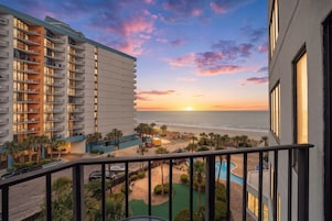 A hotel with a balcony overlooking a pool, a beach, and sunset.