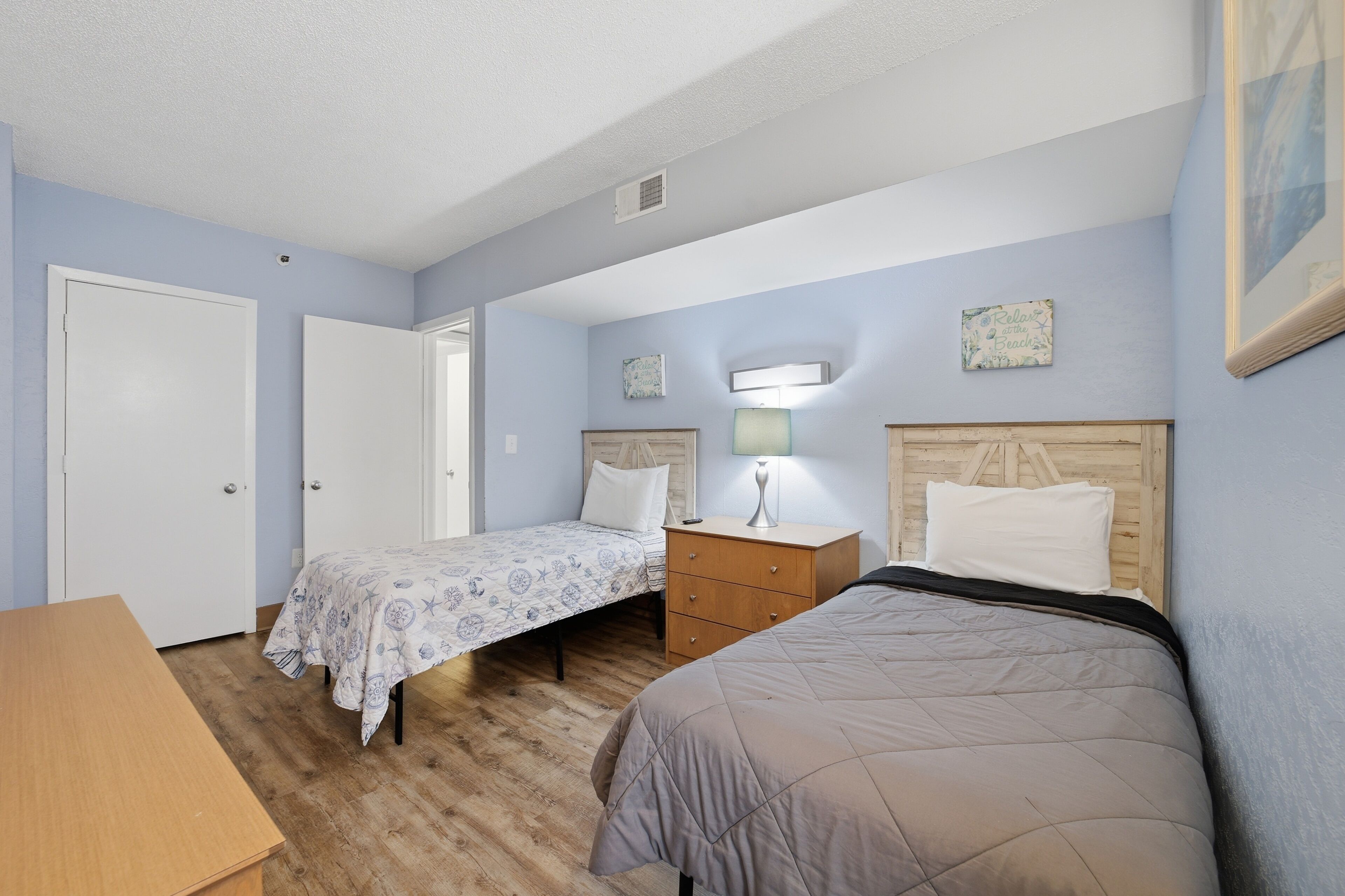A bedroom with two single beds, a wooden nightstand, a desk, and a door.