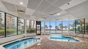 An indoor swimming pool with clear views of palm trees and a building in the background. - A Coastal Paradise - Beachfront - Gym - Pool (Myrtle Beach)