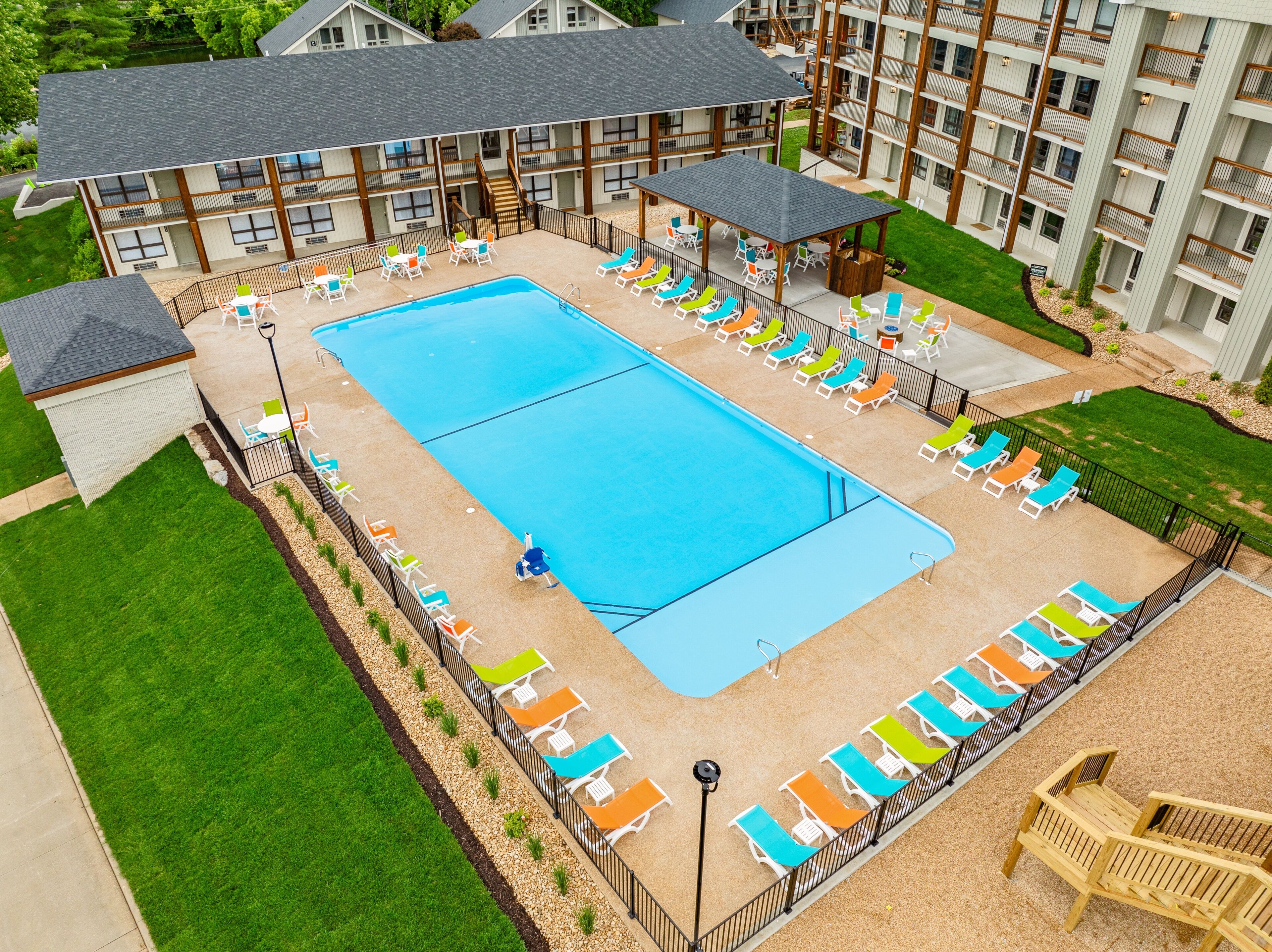 A large rectangular pool with lounge chairs and umbrellas, surrounded by a fence, with a building in the background.