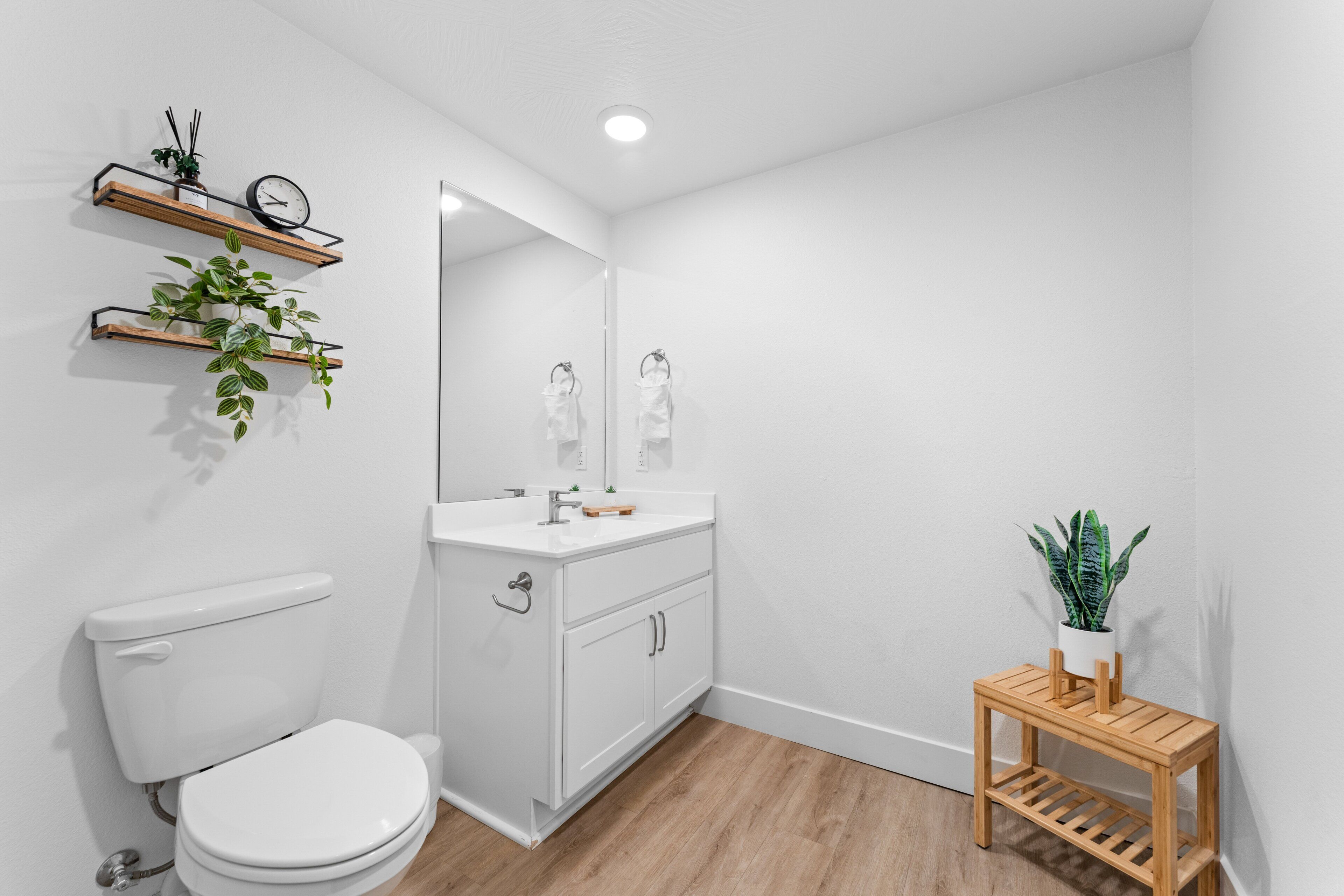 Un baño moderno con inodoro, lavabo, espejo, estantes de madera con plantas y una pequeña mesa de madera con planta en maceta.
