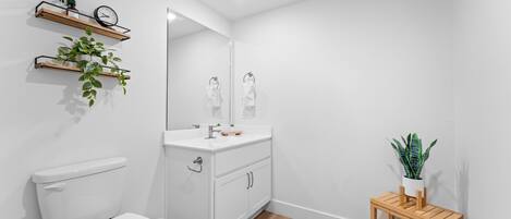 Un baño moderno con inodoro, lavabo, espejo, estantes de madera con plantas y una pequeña mesa de madera con planta en maceta.
