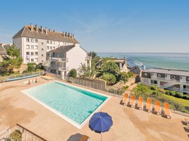 Ein Swimmingpool mit Liegestühlen und einem Sonnenschirm, umgeben von Gebäuden und Blick aufs Meer.