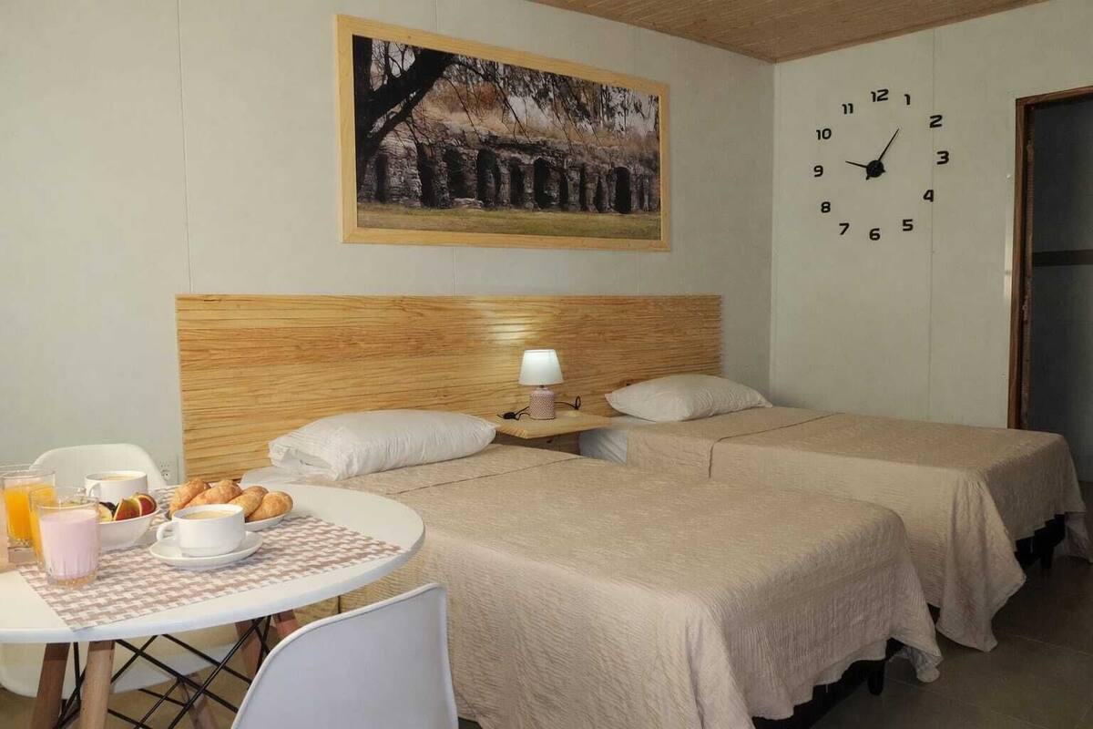A hotel room with two beds, a wooden headboard, a clock, a table with breakfast, and a painting on the wall.