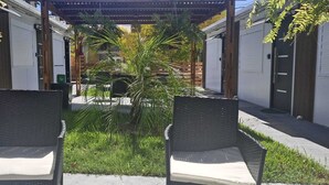 A covered outdoor seating area with two wicker chairs, a small palm tree, and a pathway leading to a building with closed doors. - Jardines Inn (Trinidad)