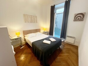 Een hotelkamer met een bed, nachtlampjes, een raam met gordijnen, een radiator en een klein kunstwerk aan de muur.