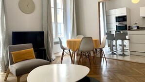 Ein modernes Wohnzimmer mit Fernseher, einem Esstisch mit Stühlen, einer Couch mit Kissen, einem großen Fenster mit Vorhängen und einer Küchenzeile mit Geräten.
