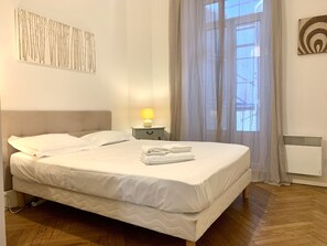 Una cama bien hecha con sábanas blancas, una lámpara de noche y una obra de arte en la pared.