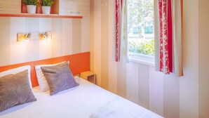 Ein Hotelzimmer mit Bett, Kissen, einem Nachttisch, einem Fenster mit Vorhängen und einem Regal mit Pflanzen.