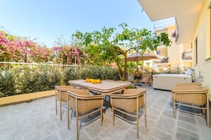 En udendørs terrasse med borde og stole, omgivet af planter og et hegn.