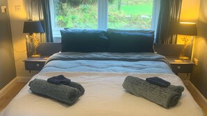 Een keurig opgemaakt bed met twee opgerolde handdoeken, een donkerblauwe hoofdbord en een raam met gordijnen.