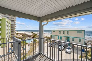 Ein Balkon mit Blick auf Strand und Meer, ein Parkplatz sowie ein Gebäude mit hellblauer Fassade.