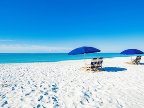 Ein Strand mit zwei blauen Sonnenschirmen und zwei Liegestühlen.