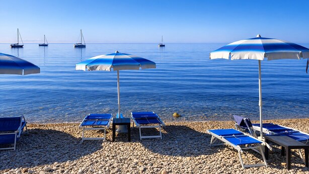 Ein Strand mit Sonnenschirmen, Liegestühlen und Booten im Wasser.