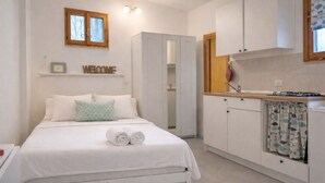 Ein Schlafzimmer mit Bett, Kissen, Nachttisch, Kleiderschrank, einer Küchenzeile mit Spüle und einem Fenster.