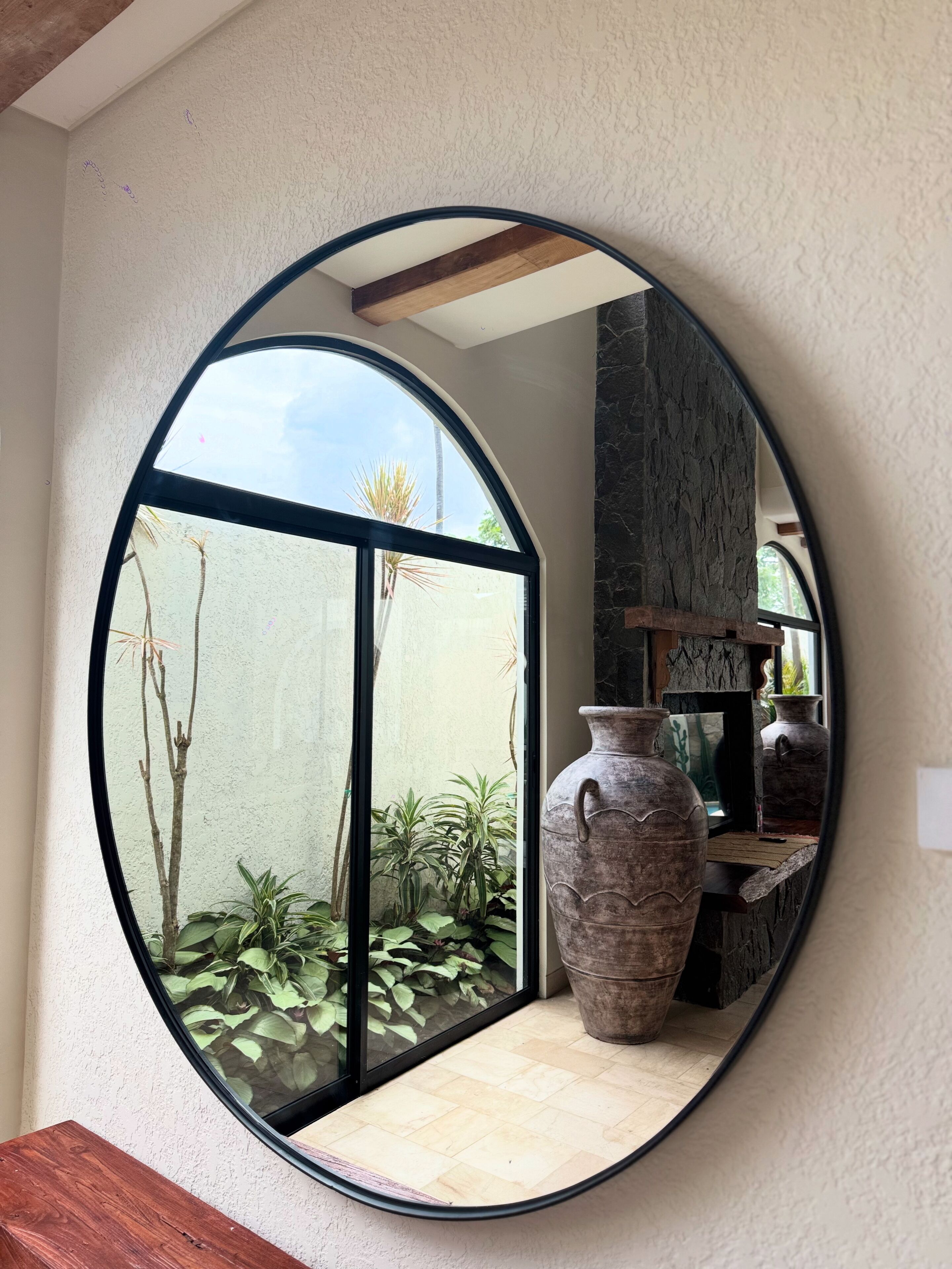 A round mirror reflecting a room with a large window, a wooden table, and a stone wall.
