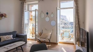 A living room with a sofa, a chair, and a table. There are large windows with a view of a body of water and a building. - L'échappée Mer (Cassis)
