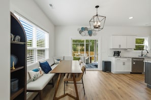 Uma cozinha moderna com armários brancos, uma área de refeições com um banco e mesa, e vista para um espaço ao ar livre através de uma porta de vidro.