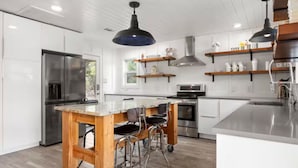 Cozinha moderna com armários brancos, geladeira de aço inoxidável, ilha de madeira com banquetas e iluminação pendente.