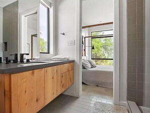 Un baño moderno con un tocador de madera, un espejo grande y vista a un dormitorio por una puerta abierta.