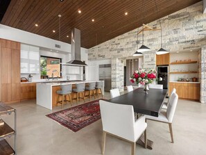 A modern kitchen with a central island, bar stools, a dining table with chairs, and a stone accent wall. - Luxury 26-Acre Compound, w/ Pool & Amazing Views (Wimberley)