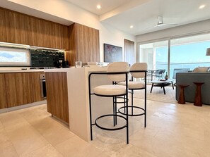 A modern kitchen with wooden cabinets, a bar area with stools, and a view of the outdoors through large windows. - Harbor 171 Unit 2209 – High-Floor Oceanview 1BR XL with Rooftop Pool (Puerto Vallarta)