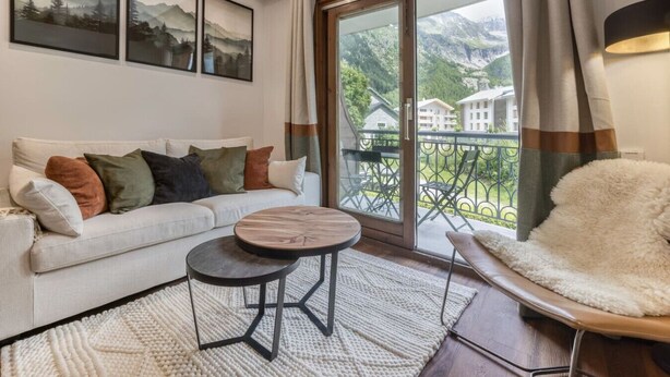 A living room with a sofa, a round table, a white furry chair, and a balcony with a view of mountains and houses. - Appart 5pax | Parking | Balconies | Views (Chamonix-Mont-Blanc)