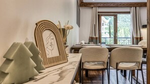 A dining area with a marble table, two chairs, a framed artwork, and a view of greenery outside. - Appart 5pax | Parking | Balconies | Views (Chamonix-Mont-Blanc)