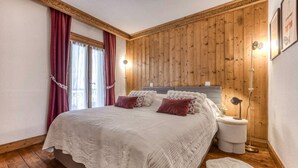A bedroom with a wooden paneled wall, a bed with a white duvet, two bedside tables with lamps, and a window with curtains. - Appart 5pax | Parking | Balconies | Views (Chamonix-Mont-Blanc)