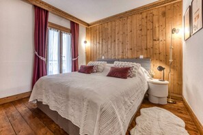 A bedroom with a wooden paneled wall, a bed with a white duvet, two bedside tables with lamps, and a window with curtains. - Appart 5pax | Parking | Balconies | Views (Chamonix-Mont-Blanc)