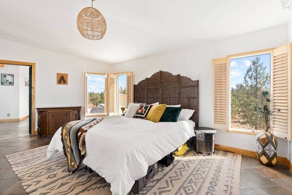 A bedroom with a large bed, a wooden headboard, a rattan pendant light, and a view of trees through the window. - Smith Rock Retreat: TWO UNITS IN ONE LISTING! (Deschutes County)