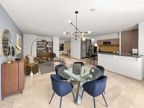 Ein modernes Wohnzimmer mit einem Glastisch, dunklen Stühlen, einer Holzkommode und einer Küche im Hintergrund.