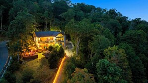 A house surrounded by dense forest with a visible driveway and a lit porch area. - 1st in Sugar/View/EV Charge/Hot Tub/Sauna/Pet Friendly/Sleeps 12/Two Master Bdrs (Sugar Mountain)