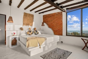 Une chambre avec un lit, une table de chevet blanche, un panier tressé suspendu, un mur en briques et une grande fenêtre donnant sur un beau paysage.