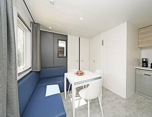 Quarto compacto com sofá azul, uma pequena mesa branca com cadeiras, cozinha compacta e janela com cortinas.